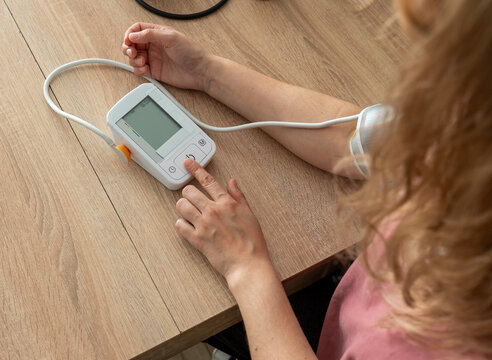 Woman starting a digital blood pressure monitor at a wooden table with a glass of water and an unlabelled white bottle nearby. Concept of home health checks and an everyday wellness routine.