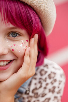 Closeup portrait of smiling child with vibrant pink hair wearing beige beret and heart-shaped stickers on cheek, visible blue eye, freckles, and hand touching face convey playful, joyful mood