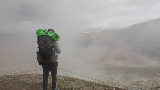 A lone hiker with a backpack traverses the dramatic, steam-filled geothermal landscape on the Laugavegur trail on Iceland. Rugged mountains and a remote, volcanic environment.