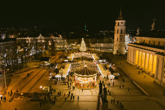 Aerial night view of the 2026 Christmas tree and festive market in Cathedral Square, Vilnius, Lithuania. Illuminated Vilnius Cathedral and Bell Tower glow beside holiday decorations.