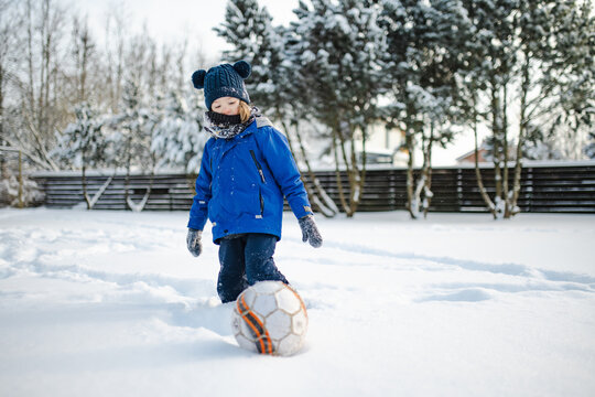 Happy young child playing with fresh snow in the backyard on a winter day. Smiling kid holding snow in gloved hands and enjoying outdoor seasonal activity.