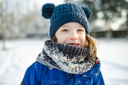 Happy young child playing with fresh snow in the backyard on a winter day. Smiling kid holding snow in gloved hands and enjoying outdoor seasonal activity.