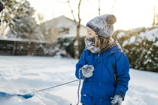 Happy young child playing with fresh snow in the backyard on a winter day. Smiling kid holding snow in gloved hands and enjoying outdoor seasonal activity.