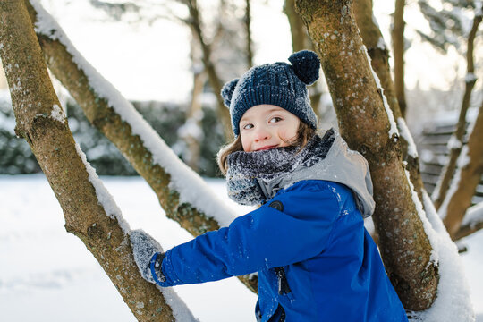 Happy young child playing with fresh snow in the backyard on a winter day. Smiling kid holding snow in gloved hands and enjoying outdoor seasonal activity.