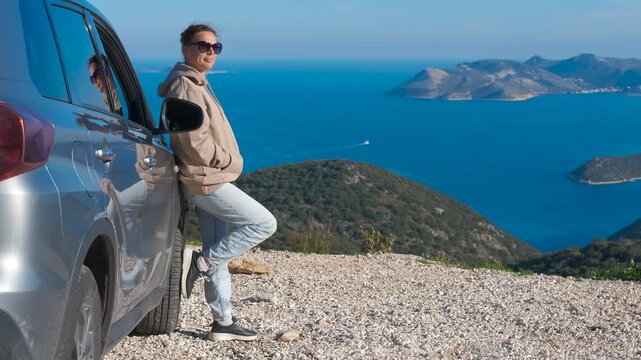 Relaxed woman admiring the beautiful seascape from a cliffside road. Young relaxed woman leaning against her car during a scenic road trip, enjoying the stunning coastal view with mountains