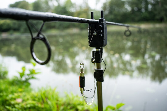 Close-up of a carp fishing rod on a bankstick with an electronic bite alarm and swinger indicator. Professional angling equipment setup near a calm lake on a rainy day.