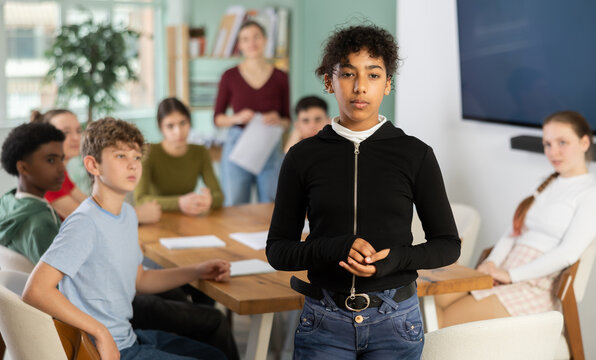 Sad teenage girl standing with her back to classmates sitting at table in classroom