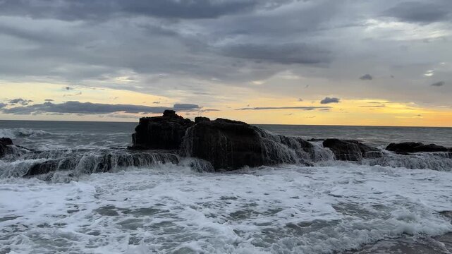 Waves hit the rocks as the sun sets over the ocean. The sky is filled with clouds and orange hues.