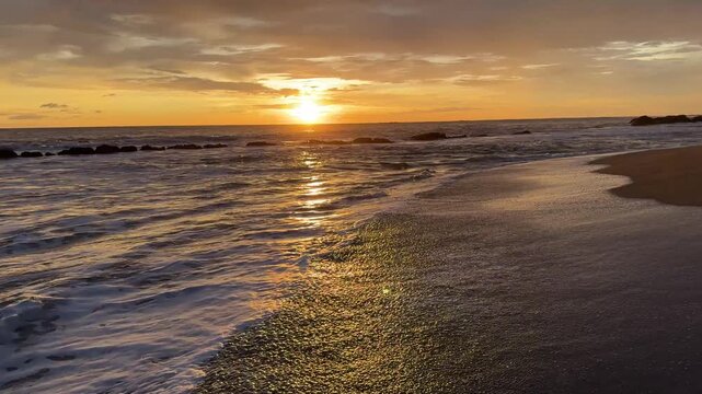Waves crash on the shore as the sun sets over the ocean. The sky displays warm colors while the water glistens in the light.
