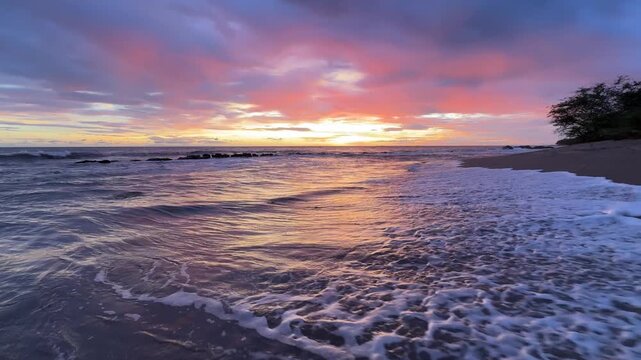 Waves hit the shore as the sun rises, casting colors in the sky at a beach. This scene shows nature's beauty at dawn.