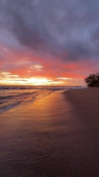 The sun sets over the ocean as waves wash onto the sandy beach. The sky shows bright colors in the evening light near a shoreline.