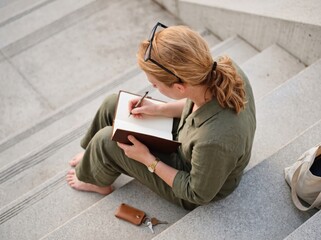 Obraz premium Focused Woman Sitting on Urban Steps Writing Personal Goals and Plans in a Notebook