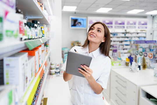 Smiling female medical worker holding a digital notepad while writing notes and managing supplies in a modern pharmacy retail store.
