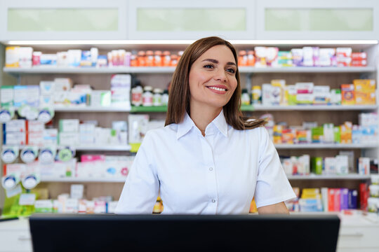 A friendly medical professional standing behind a counter inside a pharmacy surrounded by shelves stocked with pharmaceutical products and medications.