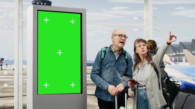 Chroma key departure board stands beside an elderly couple at the airport as they plan an itinerary and travel agenda, highlighting unconventional lifestyle, world travel and green screen use.