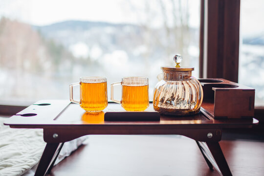 Drinking hot tea enjoying winter landscape in mountains. Close up of two cups and teapot on tray by big window of cabin house 
