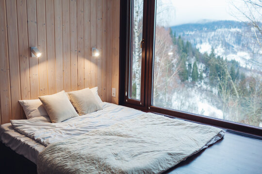 Interior of cabin house with winter landscape in mountains. Bedroom with panoramic window and snowy forest view