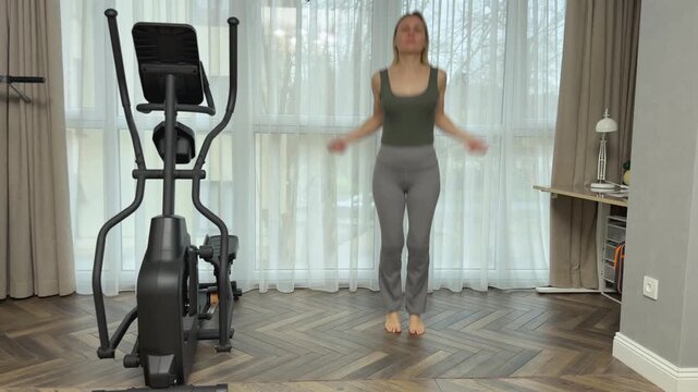 Woman in a dark sports top and light gray fitted leggings performs jump rope imitation without equipment in a bright living room with panoramic curtains, wooden floor and elliptical machine nearby