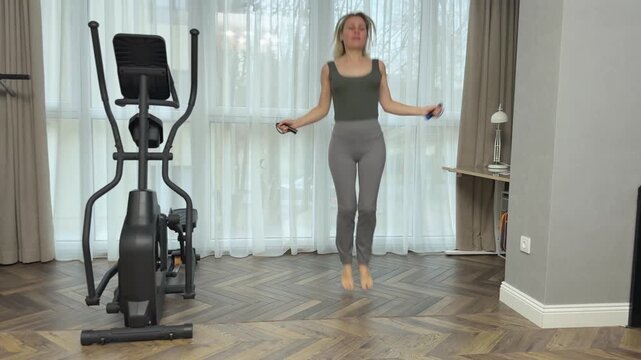Woman in dark fitted sports top and light gray leggings jumps rope barefoot in a spacious living room with herringbone floor, large sheer curtains and an elliptical trainer in the background. Home