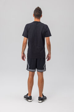 Studio photo: Back view of a muscular male model wearing a plain black cotton t-shirt and athletic shorts with white stripe details. Isolated on grey.