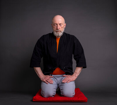 portrait of bald and bearded senior man wearing a short kimono and sitting in a traditional Japanese seiza position on a cushion pad, contemplating, meditating or praying concept