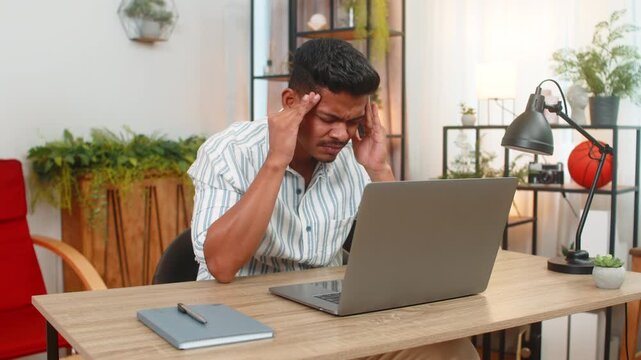 Indian man at home feels headache during laptop work rubs temples squints at screen and slows pace. Arabian guy at table breathes deep drinks water dims brightness and plans short break for calm rest