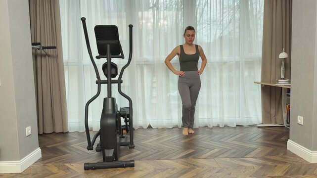 Woman in fitted sportswear top, leggings and sneakers performs deep forward lunges in bright living room near elliptical trainer. Home workout for legs strength, balance and mobility.