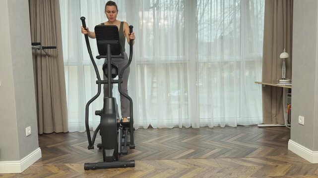 Woman performs reverse stepping on elliptical machine using moving handles. Balanced home workout engaging different muscle groups.