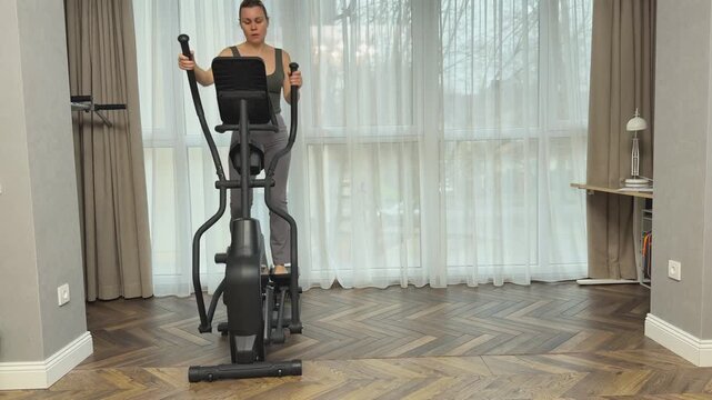 Woman steps forward on elliptical machine synchronizing arm and leg movement. Effective home cardio session in modern living room.