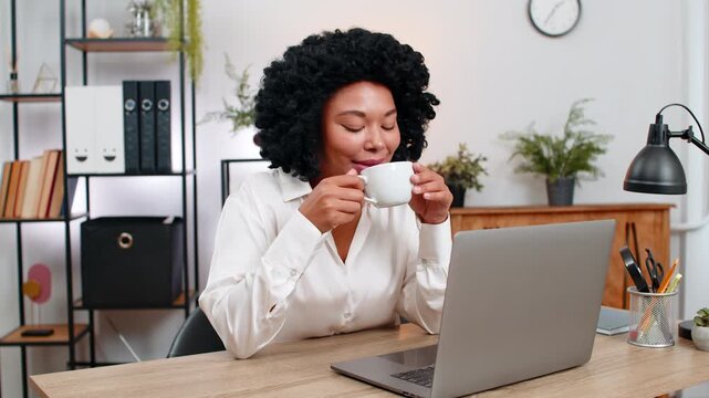 African American businesswoman at home office slowly sips coffee gazing out window letting ideas flow. Freelancer girl at table pauses work enjoying calm drink moment relaxing mind before next task.