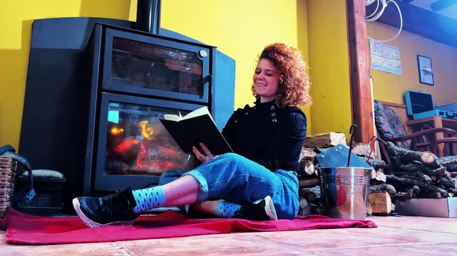 Young woman relaxing and reading a book by the warm fireplace in a cozy rural home