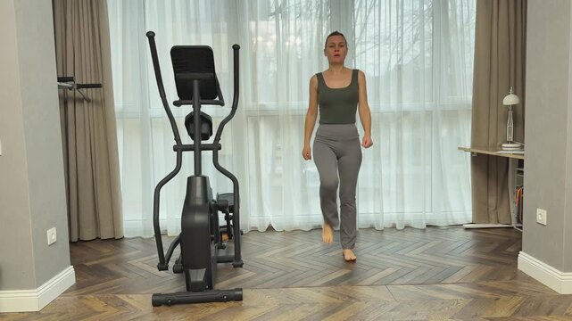 Woman in sports top and leggings walks calmly in place with arms lifted overhead. Light recovery movement in living room after cardio workout.