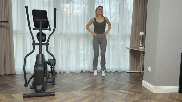 Woman in sports top and leggings marches in place lifting knees high with hands on hips. Simple home warmup exercise.