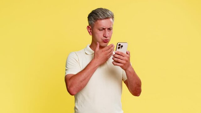 Middle-aged man staring at smartphone with hopeless expression, covering face with hand in despair after receiving bad news or losing money. Young guy showing failure emotion on yellow background