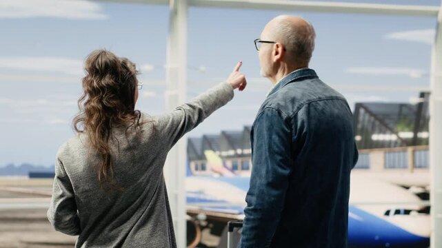 Joyful elderly couple points toward the runway while waiting in a priority boarding queue at the terminal, with luggage ready for departure.