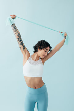 Slim woman in sportswear practicing with a resistance band over a blue backdrop, focusing on stretching