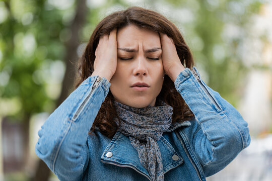Young woman with headache walking in city street holds head and feels pain during sudden migraine attack. Girl tourist suffering outdoors shows discomfort tension and fatigue during urban park walk.
