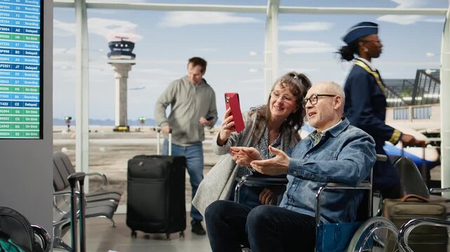 Older man in a wheelchair takes photos with his wife in an airport lounge while waiting for priority boarding and enjoying retirement adventure.