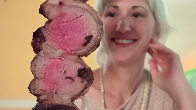 Woman is happily looking at traditional Brazilian picanha roast beef skewer with grilled, juicy, medium-rare slices of meat, enjoying delicious churrascaria experience. Elegant luxury romantic dinner.