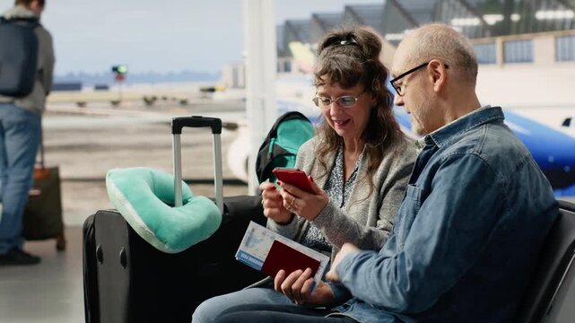Confident senior couple using smartphone to check travel itinerary and sightseeing list, sitting in airport terminal, highlighting unconventional lifestyle.