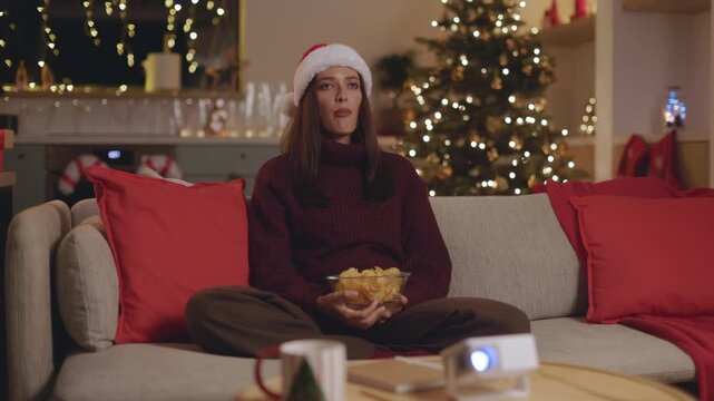 Bored woman in Santa hat eating chips, watching movie at home, 4K