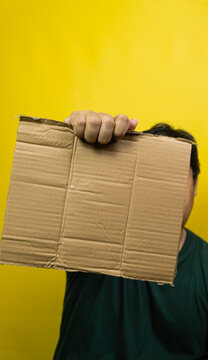 An Asian man holding a blank piece of brown corrugated cardboard in front of the right side of his face against a solid yellow background. The left side of the head and ear are visible