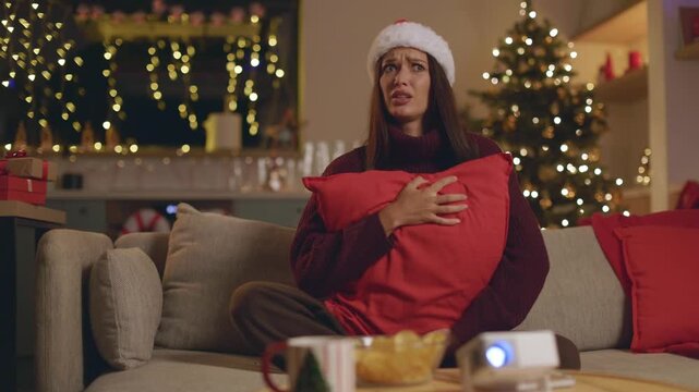 Frightened woman in Santa hat hiding behind pillow while watching scary movie
