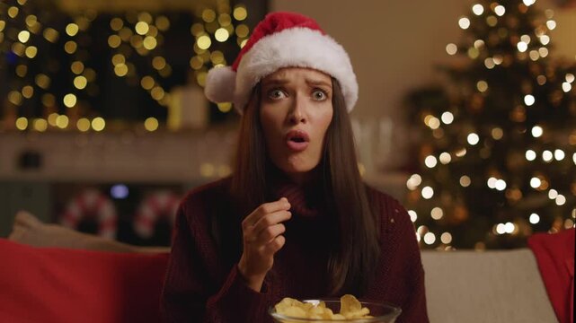 Worried young woman in Santa hat staring wide-eyed and eating chips, 4K