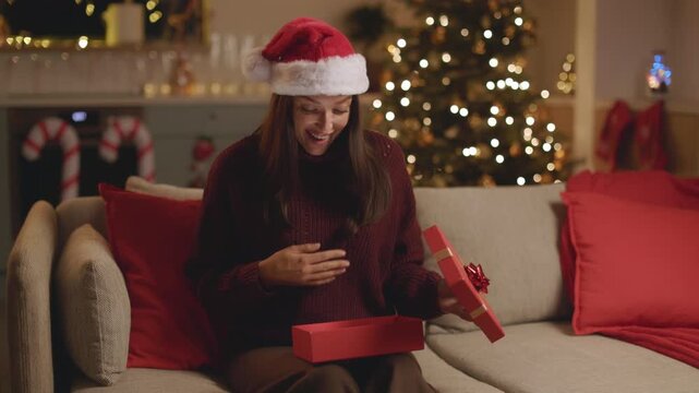 Smiling woman in Santa hat opening red gift box on couch, 4K