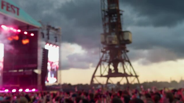 Blurred outdoor concert crowd with stage lights, pink smoke, towering rigging, and dramatic cloudy sky at sunset