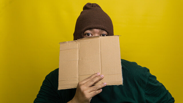 Young Asian male wearing brown balaclava obscures lower visage with blank corrugated board against vivid yellow backdrop. Only observant eyes remain visible above textured placard,