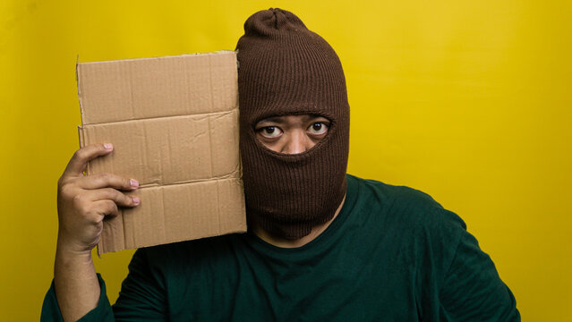Young Asian male wearing brown balaclava holds blank corrugated board next to head against vibrant yellow backdrop. The masked individual stares directly forward, clad in dark green