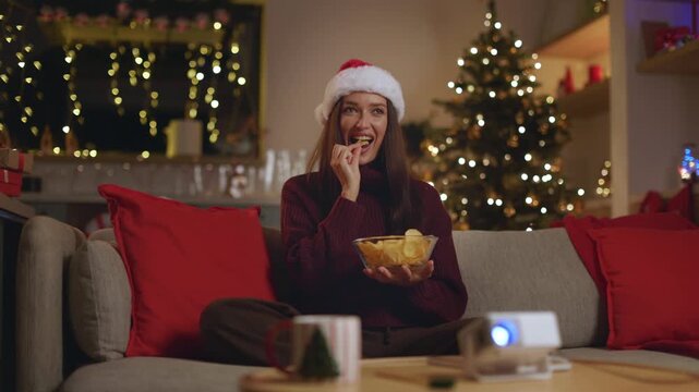Woman in Santa hat eating potato chis and watching movie with projector
