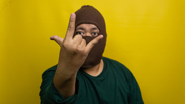 Young Asian male wearing brown balaclava displays rock and roll hand gesture against vivid yellow backdrop. The masked individual extends index and pinky fingers while holding thumb